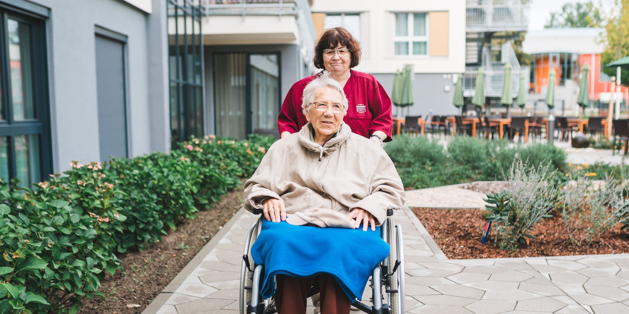 Staatlich anerkannte*r Krankenpflege-/ Altenpflegehelfer*in schiebt Bewohnerin im Rollstuhl durch den Garten