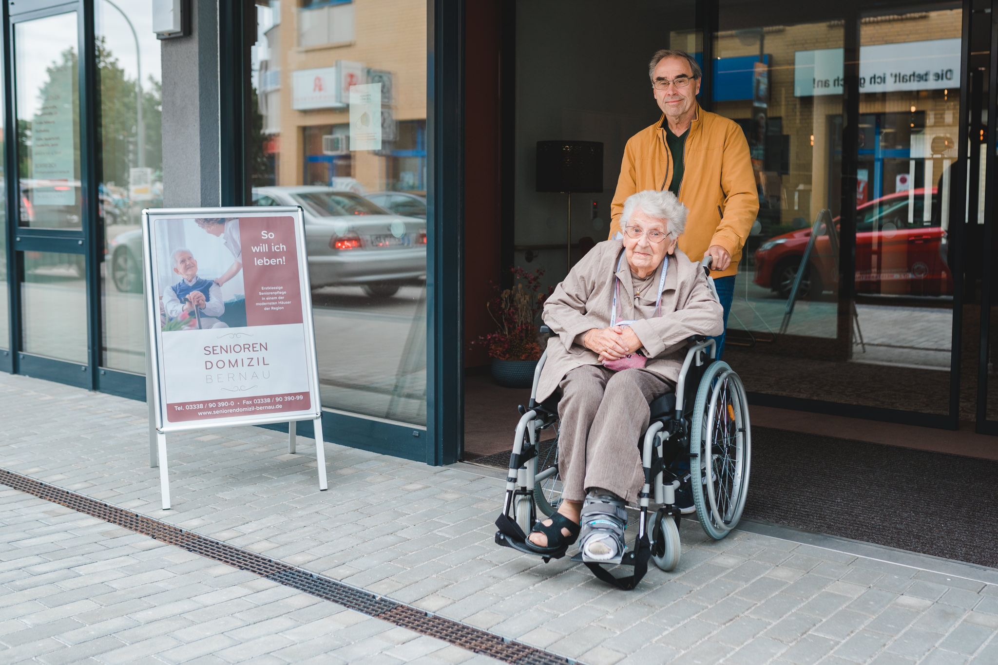 Angehöriger schiebt pflegebedürftige Mutter mit Rollstuhl aus Seniorendomizil